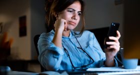 Confused young woman designer sitting in office working using mobile phone at night.